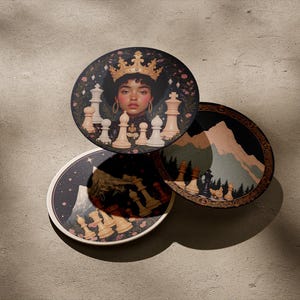 Puede incluir: Tres posavasos redondos con fondo negro y un diseño de piezas de ajedrez blancas. El posavasos superior presenta un retrato de una mujer con una corona dorada y rodeada de piezas de ajedrez. Los otros dos posavasos presentan una escena de montaña con piezas de ajedrez en primer plano.