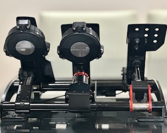 May include: Black racing simulator pedals with silver accents and red details. The set includes two circular units and a pedal on the right. The brand name is visible on the circular units. The pedals are mounted on a black frame.