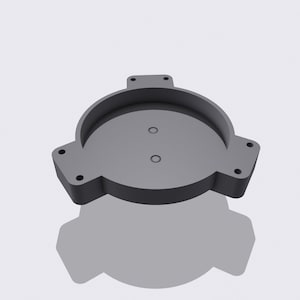 Puede incluir: Un objeto gris circular impreso en 3D con un borde elevado y tres pestañas rectangulares. Dos pequeños agujeros son visibles en el centro. El objeto está sobre un fondo blanco.