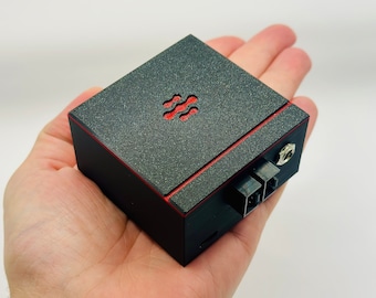 May include: A small, black electronic device with a textured, dark gray top and red accents. The top features a red, stylized wave symbol. The device has ports and a jack on the side and is held in a hand.