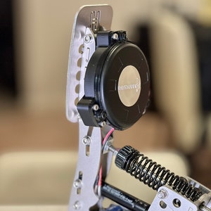 Puede incluir: Primer plano de un pedal de carreras plateado y negro con un resorte y un motor negro con el texto "HYSTERONIC" en él.