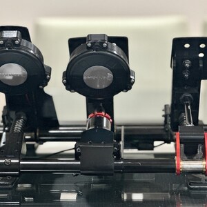 May include: Black racing simulator pedals with a metal frame and red accents. The pedals have a spring-loaded design and are adjustable. The pedals are labeled "Simucube".