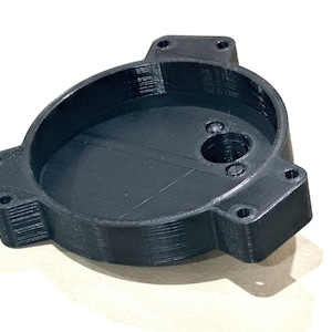 Puede incluir: Una pieza de plástico negro impresa en 3D con una abertura circular y varios agujeros. La pieza tiene una superficie lisa y redondeada y una apariencia texturizada y en capas.