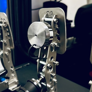 May include: Close-up of a silver metal racing pedal set with a black background. The pedals have a textured surface and are connected by a metal bar. The pedals are designed for use with a racing simulator.