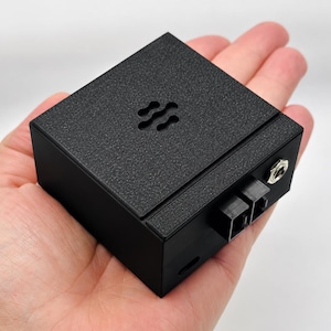 May include: A compact, matte black square electronic device with a textured top surface featuring a small, wave-like cutout pattern. The side panel includes two black square ports and a single circular metallic audio jack input port.