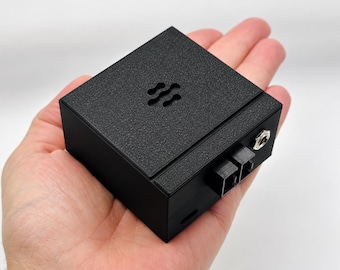 May include: A compact, matte black square electronic device with a textured top surface featuring a small, wave-like cutout pattern. The side panel includes two black square ports and a single circular metallic audio jack input port.