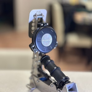 Puede incluir: Un pedal de batería plateado y negro con un reposapiés negro y un resorte plateado. El pedal tiene un mando de control redondo negro con el texto "Dayton Audio" impreso en él.