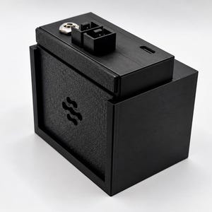Puede incluir: Una carcasa rectangular negra para dispositivos electrónicos con un panel frontal texturizado que presenta un pequeño recorte de logotipo. La superficie superior incluye un puerto circular metálico, un conector rectangular negro y una pequeña ranura de entrada.