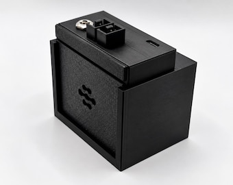 Könnte beinhalten: Ein schwarzes, rechteckiges Gehäuse für elektronische Geräte mit einer texturierten Frontplatte, die einen kleinen Logo-Ausschnitt aufweist. Die Oberseite enthält einen metallischen runden Anschluss, einen schwarzen rechteckigen Stecker und einen kleinen rechteckigen Eingangsschlitz.