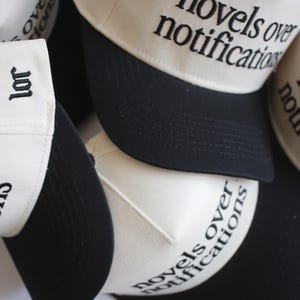 May include: A collection of white and black baseball caps. The caps have a white crown and a black brim. The phrase "novels over notifications" is embroidered on the front of the caps in black lettering. Some caps have the word "lor" embroidered on the side.