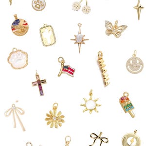 Op de afbeelding: Een verzameling goudkleurige bedels in verschillende vormen, waaronder een hart met de tekst "honey", een kruis met een edelsteen, een ster, een vlinder en een smiley. Andere bedels zijn een cactus, een motorfiets en een voetbal.