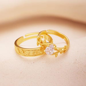 May include: A pair of gold rings with a delicate floral design. The larger ring features a small, clear heart-shaped stone.