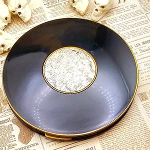 May include: A black compact with a gold rim and a silver floral design in the center. The compact is closed and sitting on a newspaper.