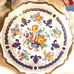 May include: A round porcelain plate with a scalloped edge and gold trim. The center features a colorful floral bouquet with pink, orange, and blue flowers. A dark blue decorative border with gold filigree accents surrounds the central design.
