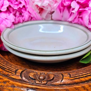 May include: Two white ceramic bowls with a green trim are stacked on a wooden decorative tray. The bowls are surrounded by pink peonies. The tray has a carved floral design.