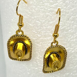 May include: A pair of gold-tone earrings featuring a cowboy hat design. The hats have a textured surface and a beaded trim.
