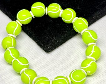 May include: A circular bracelet composed of round, neon yellow beads, each featuring a white curved line pattern that mimics the appearance of a tennis ball. The beads are strung together in a continuous loop, creating a vibrant, sporty accessory.