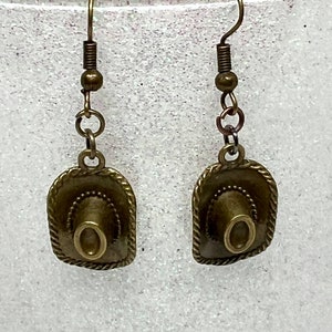 May include: A pair of bronze-colored earrings featuring a miniature cowboy hat charm. The hats have a detailed design and are attached to a simple hook.