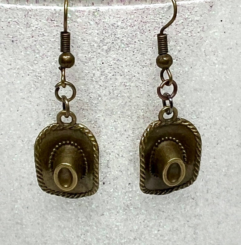 May include: A pair of bronze-colored earrings featuring a miniature cowboy hat charm. The hats have a detailed design and are attached to a simple hook.