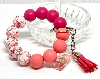May include: A pink and white silicone beaded bracelet with a floral pattern and a pink tassel keychain. The bracelet has a silver metal clasp and a silver chain attached to the tassel.