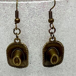 May include: A pair of bronze-colored earrings featuring a miniature cowboy hat charm. The hats have a detailed design and are attached to a simple hook.