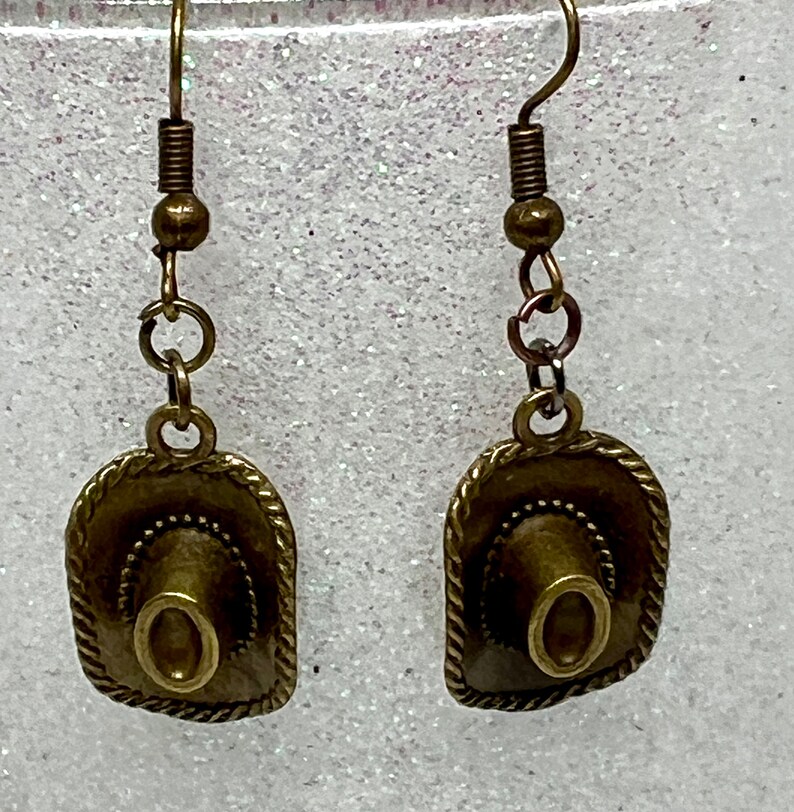 May include: A pair of bronze-colored earrings featuring a miniature cowboy hat charm. The hats have a detailed design and are attached to a simple hook.