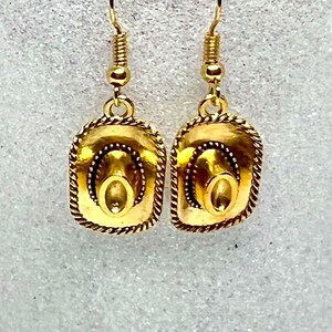May include: A pair of gold-tone dangle earrings featuring a cowboy hat design. The hats have a rope-like border and a detailed design.