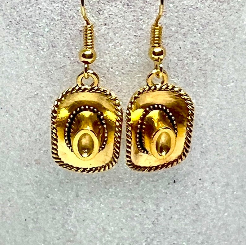 May include: A pair of gold-tone dangle earrings featuring a cowboy hat design. The hats have a rope-like border and a detailed design.