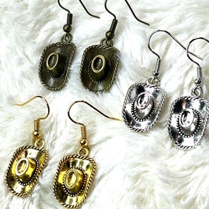 May include: Four pairs of gold and silver earrings with a cowboy hat design. The earrings are on a white fuzzy surface.