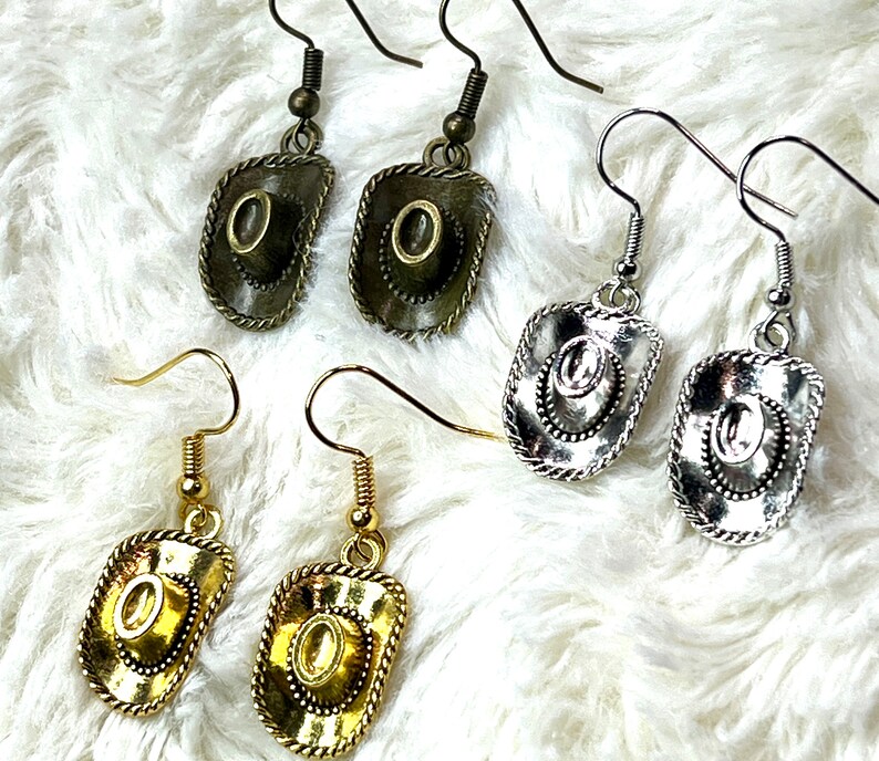 May include: Four pairs of gold and silver earrings with a cowboy hat design. The earrings are on a white fuzzy surface.