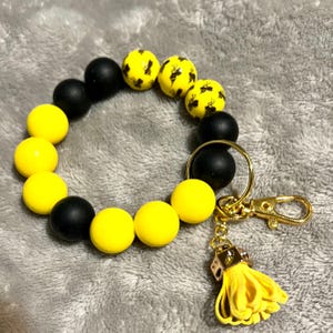 May include: A black and yellow beaded bracelet with a gold key chain and tassel. The beads are round and have a matte finish. The keychain has a small gold charm in the shape of a dice.