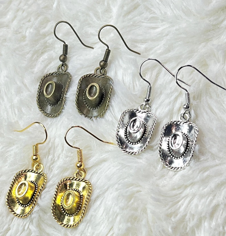 May include: Three pairs of dangle earrings with a cowboy hat charm. The earrings are available in gold, silver, and bronze.
