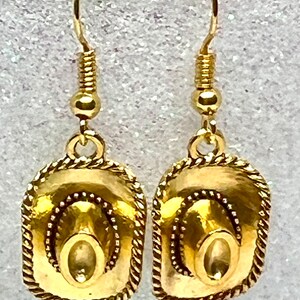 May include: Gold-tone dangle earrings featuring a cowboy hat design. The hats have a rope-like border and are detailed with a raised center.