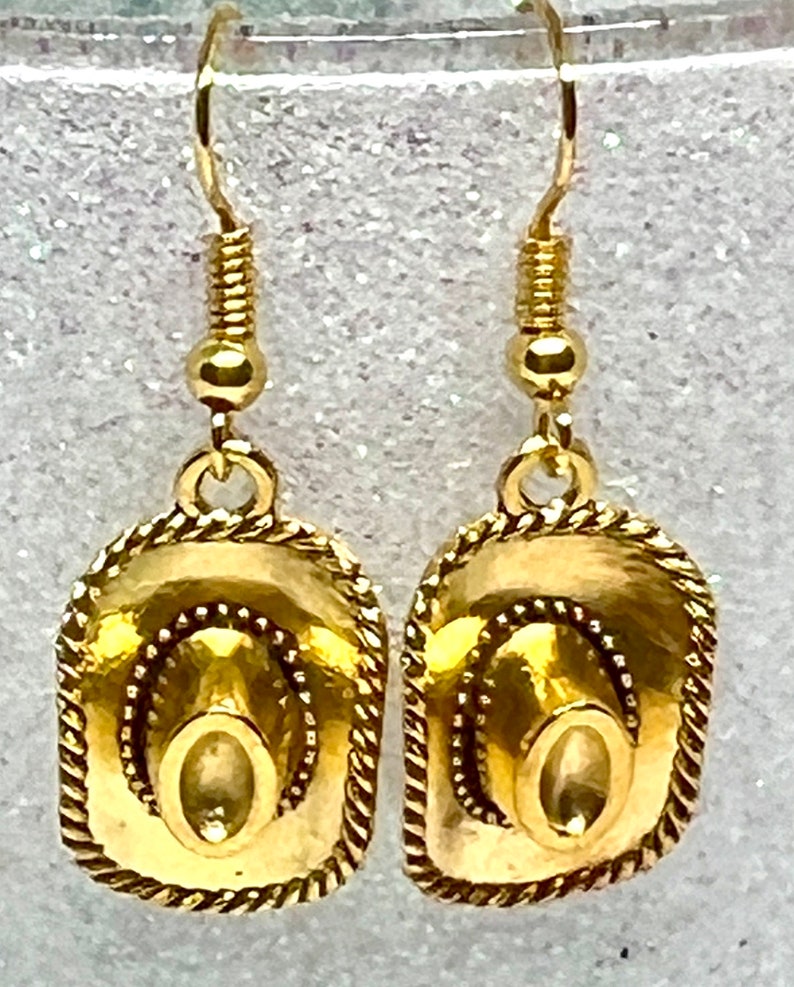 May include: Gold-tone dangle earrings featuring a cowboy hat design. The hats have a rope-like border and are detailed with a raised center.