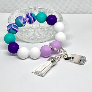 May include: A colorful silicone beaded bracelet with a white tassel and a charm of a boba tea cup. The bracelet is made of white, purple, turquoise, and a swirl pattern of purple, white, and turquoise beads.