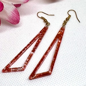 May include: A pair of dangle earrings with a geometric, triangular shape. The earrings are made of clear resin with a copper glitter embedded in the resin. The earrings have gold hooks.
