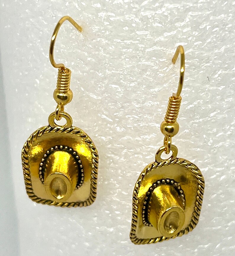 May include: A pair of gold-tone earrings featuring a cowboy hat design. The hats have a textured surface and a beaded trim.