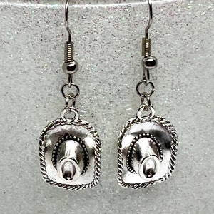 May include: Silver dangle earrings featuring a miniature cowboy hat charm. The hats have a detailed design with a rope trim.