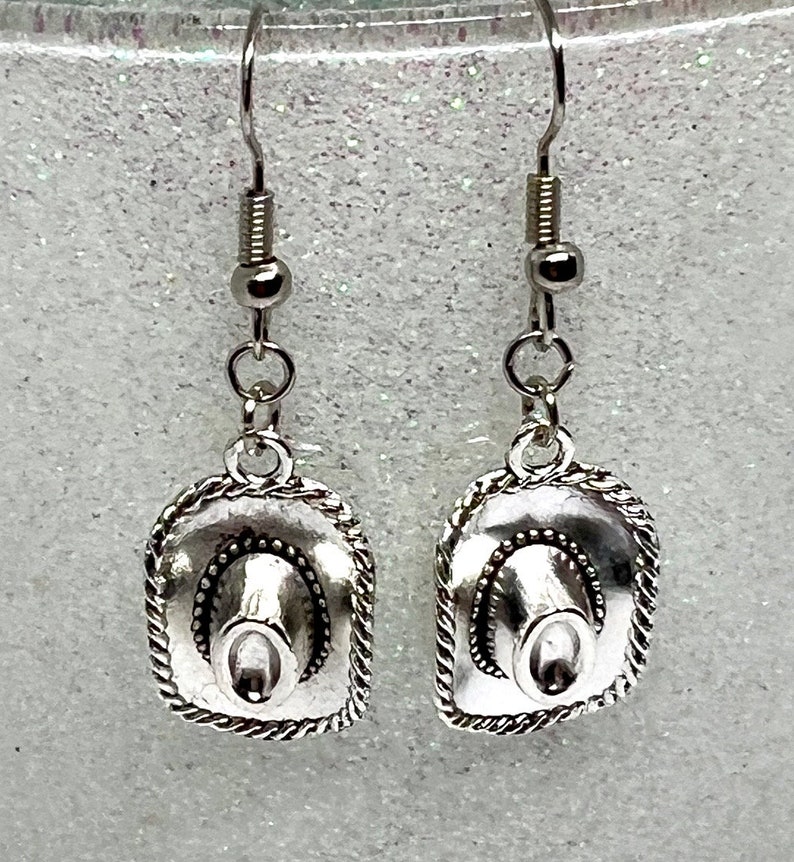 May include: Silver dangle earrings featuring a miniature cowboy hat charm. The hats have a detailed design with a rope trim.