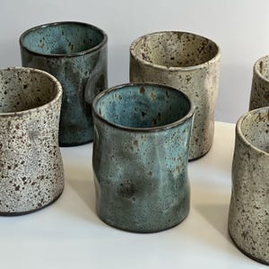 Op de afbeelding: Set van zes keramische mokken met een gespikkeld glazuur in tinten bruin, wit en blauw. De mokken hebben een unieke, licht onregelmatige vorm.