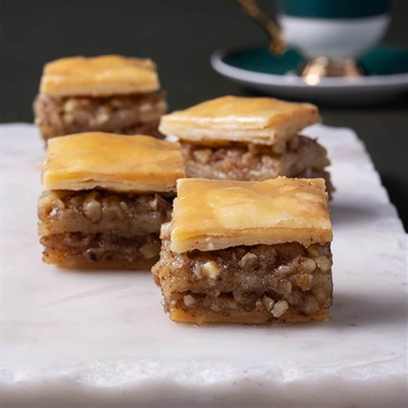 May include: Four pieces of baklava, a traditional Middle Eastern dessert, are arranged on a white marble surface. The baklava is made with layers of flaky pastry, filled with chopped nuts, and drizzled with honey or syrup.