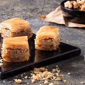 May include: Three pieces of baklava on a black marble serving tray. The baklava is made with layers of phyllo dough, filled with chopped nuts, and drizzled with honey syrup. The tray is surrounded by chopped walnuts.