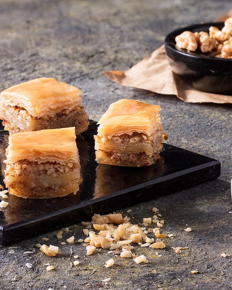 May include: Three pieces of baklava on a black marble serving tray. The baklava is made with layers of phyllo dough, filled with chopped nuts, and drizzled with honey syrup. The tray is surrounded by chopped walnuts.