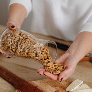 May include: A person's hand pouring walnuts from a glass jar onto a wooden cutting board with slices of brie cheese.
