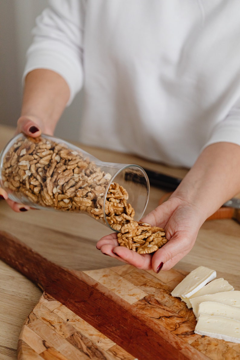 May include: A person's hand pouring walnuts from a glass jar onto a wooden cutting board with slices of brie cheese.