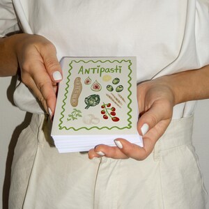 Puede incluir: Una libreta blanca con un borde verde y la palabra "Antipasti" en letras verdes. La libreta tiene ilustraciones de varios ingredientes de antipasti italianos, como pan, alcachofas, higos, aceitunas y tomates.