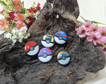 May include: Six colorful round buttons with different designs, including a rainbow, a yellow and black design, a red and white design, and a blue and white design. The buttons are on a brown wooden surface.