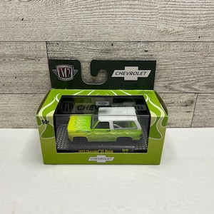 May include: A die-cast 1973 Chevrolet K5 Blazer model in a green and white color scheme, with flame graphics. The model is displayed in a green and yellow box with the Chevrolet logo.