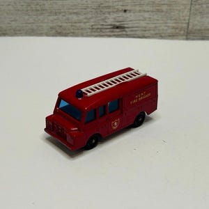 May include: A red die-cast toy fire engine featuring a white ladder mounted on the roof, blue windows, and black wheels. The side of the vehicle displays the text Kent Fire Brigade in yellow lettering along with a small red and white shield emblem.