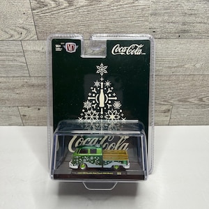 May include: A green and white 1957 VW Double Cab Truck USA Model toy truck, with a wooden bed, displayed in a clear plastic case. The packaging features the Coca-Cola logo and a Christmas tree design.